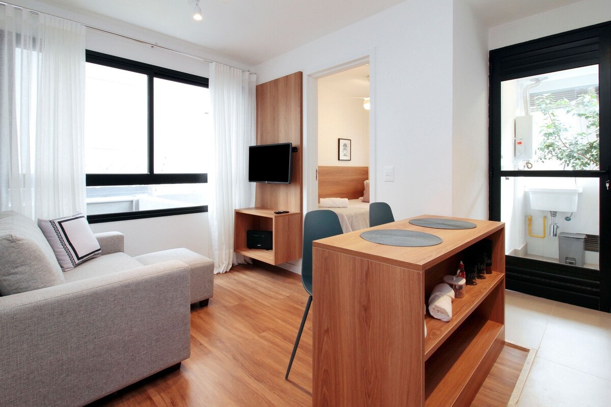 A modern hotel room with a sofa, a TV, a wooden desk with a flat-screen TV, a bed, and a bathroom visible through a glass door.