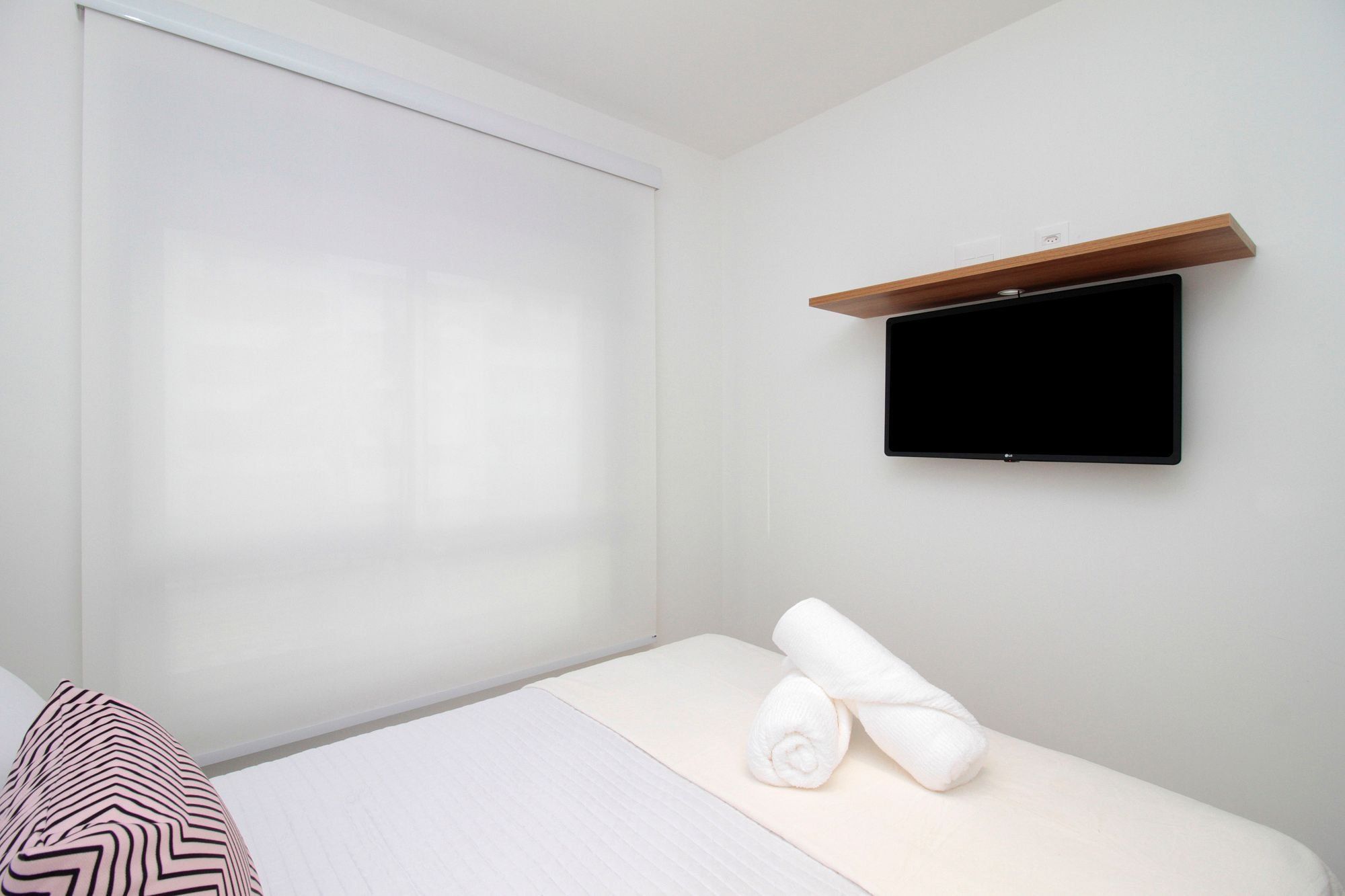 Habitación de hotel con cama, televisión montada en la pared y ventana con persianas.
