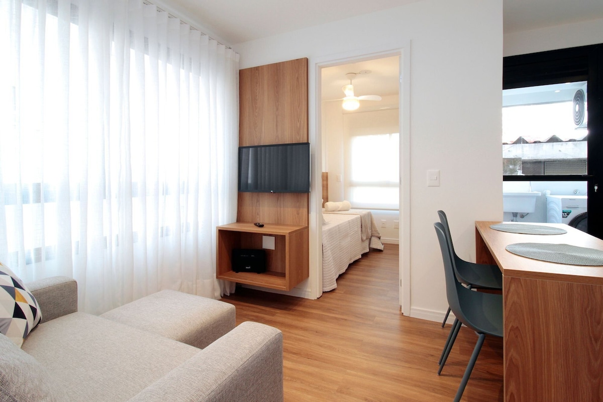 A modern living room with a sofa, a TV mounted on a wooden panel, a dining area with a table and chairs, and a view of the outside through a window.
