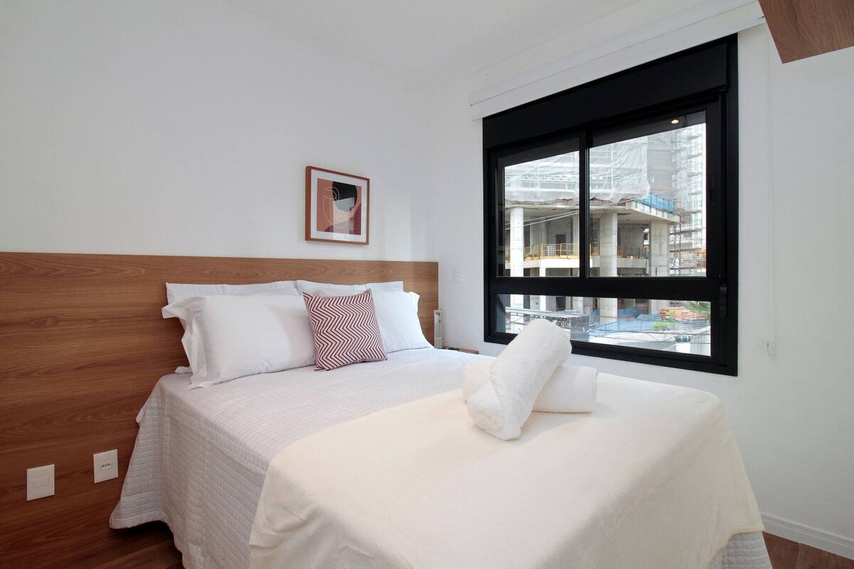 A hotel room with a wooden headboard, a large window with a view of a building under construction, and a neatly made bed with white linens.