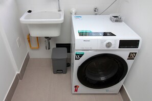 A compact laundry room with a washing machine, a small sink, and a trash bin.