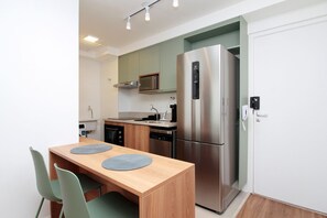 A compact kitchen with a stainless steel refrigerator, a microwave, and a dishwasher. There is a wooden dining table with two chairs and two placemats. - Boulevard Agami (São Paulo)
