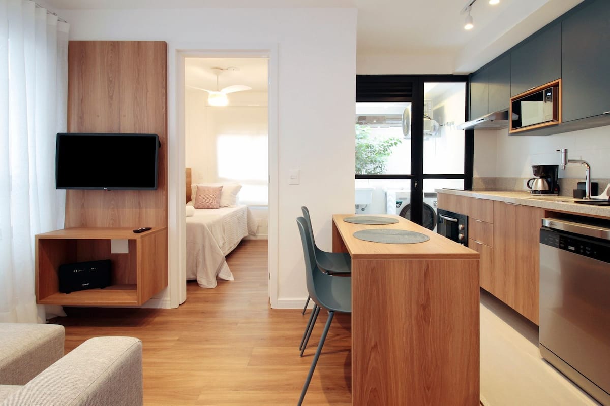 A modern hotel room with a TV, wooden furniture, a dining area, and a kitchen.