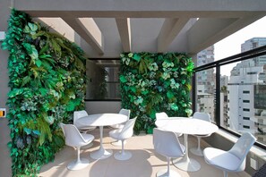 A balcony with white round tables and chairs, surrounded by lush green foliage. - Boulevard Agami (São Paulo)