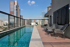 Una piscina en la azotea con agua cristalina y azul, rodeada de edificios altos, y amueblada con sillas de mimbre.
