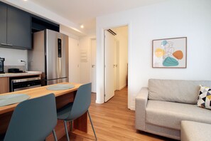 A modern kitchen with a built-in refrigerator, a dining area with a wooden table and chairs, and a living room with a sofa and a wall-mounted abstract painting. - Boulevard Agami (São Paulo)