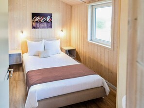 Un dormitorio con pared de paneles de madera, una cama con ropa blanca, dos mesitas de noche y una ventana con vistas.