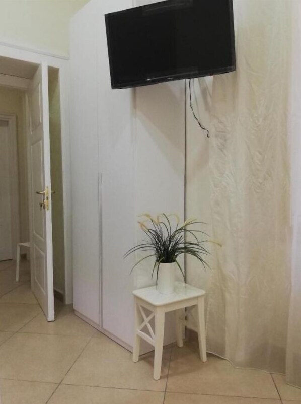 Una habitación con un armario blanco, una mesita pequeña con una planta y un televisor montado en la pared.