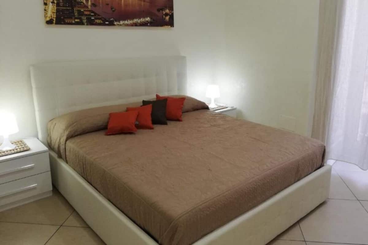Un lit bien fait, avec une couvre-lit beige et des coussins rouges et noirs, une table de chevet blanche avec une lampe, et une commode blanche.