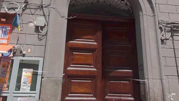 Un edificio con una amplia puerta doble de madera, un pequeño balcón con barandilla de metal y una ventana con contraventanas verdes.
