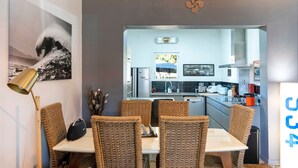 A dining area with wicker chairs, a white table, a modern kitchen in the background, and a wall-mounted light fixture. - Vacation Home with Garden in Hossegor, Near Lake, Town Center & Beaches – Perfect for 4-6 Guests (HOSSEGOR)