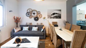 A modern living room with a sofa, dining table, wicker chairs, wall decorations, and a ceiling light fixture. - Vacation Home with Garden in Hossegor, Near Lake, Town Center & Beaches – Perfect for 4-6 Guests (HOSSEGOR)