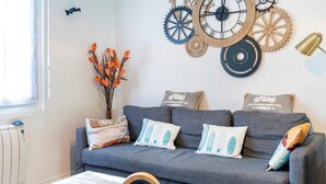 A living room with a grey sofa, decorative pillows, a wall clock, and a vase with orange flowers. - Vacation Home with Garden in Hossegor, Near Lake, Town Center & Beaches – Perfect for 4-6 Guests (HOSSEGOR)