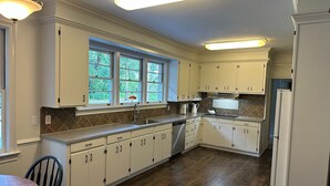 Una cocina con armarios blancos, piso de madera y ventana con vista a follaje.