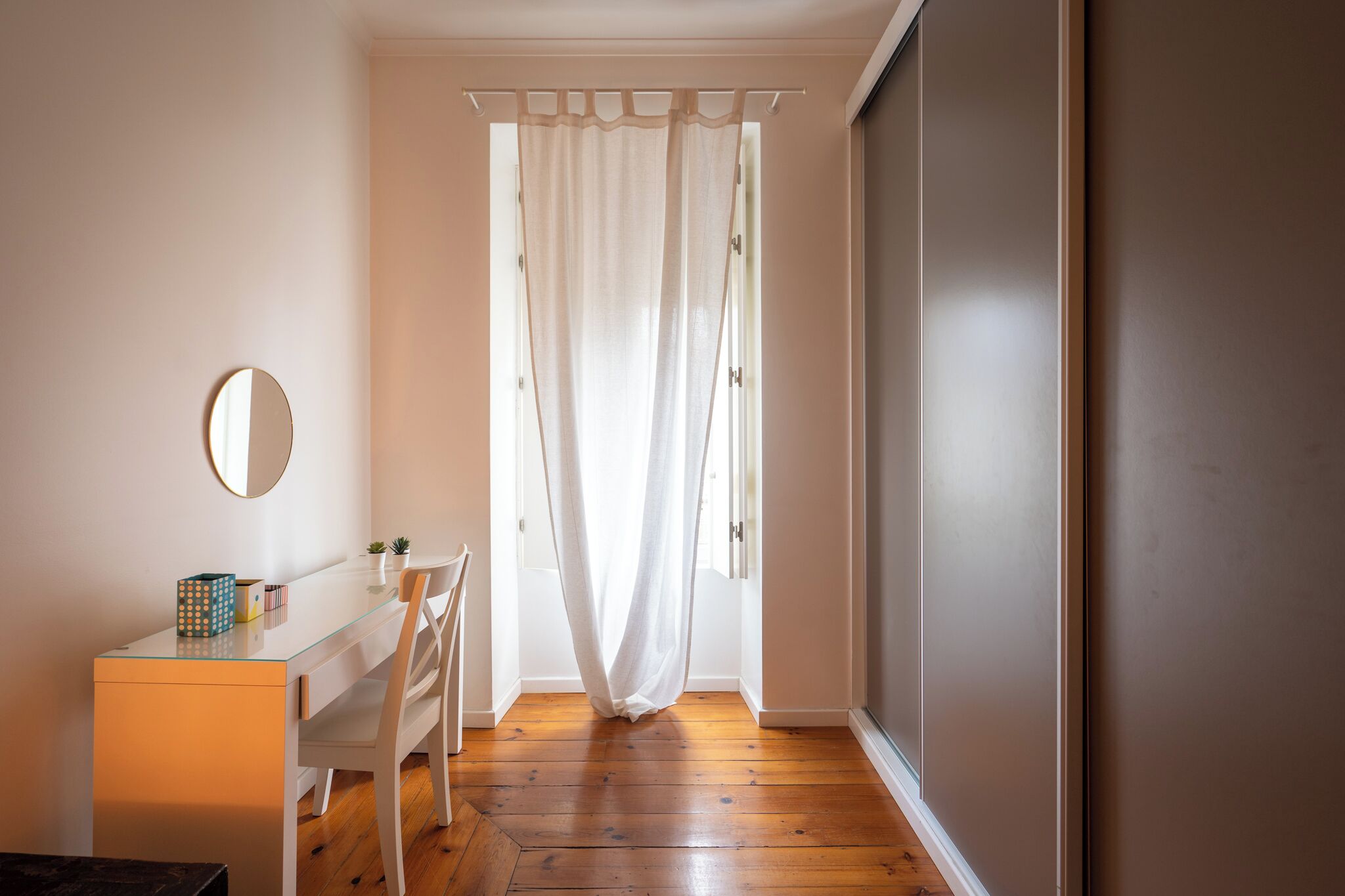 Una habitación con un escritorio de vidrio, un espejo redondo, una silla blanca, una planta pequeña y una cortina blanca.