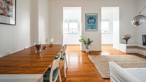 A modern living room with a wooden dining table, a sofa, and a TV stand. There are two windows providing natural light and a framed picture on the wall.