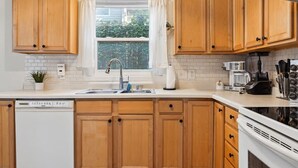 Una cocina con armarios de madera, encimera blanca, lavavajillas, fregadero, ventana con cortinas y estufa.