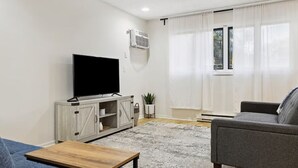 Una sala de estar con un televisor de pantalla plana sobre un mueble de madera, un sofá gris, una mesa de centro de madera y un ventanal con cortinas de gasa.