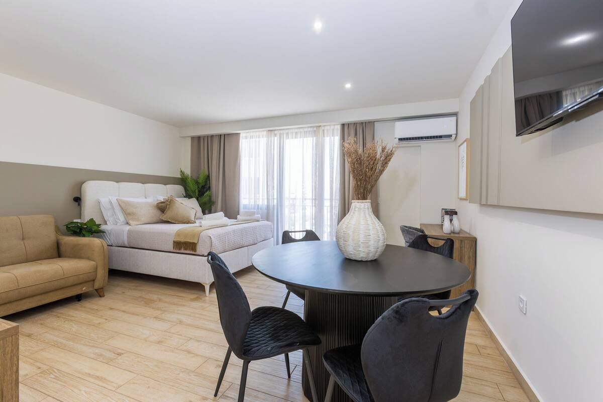 Un salon moderne comprenant un canapé, une table à manger avec des chaises, un téléviseur fixé au mur et une grande fenêtre avec des rideaux.