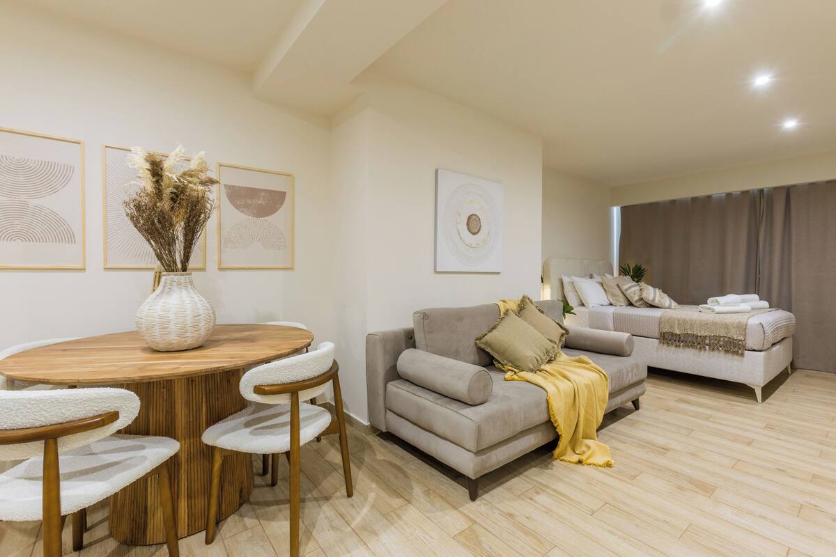 Un salon moderne avec une table ronde en bois, deux chaises blanches, un canapé gris et un lit recouvert d’un plaid jaune.