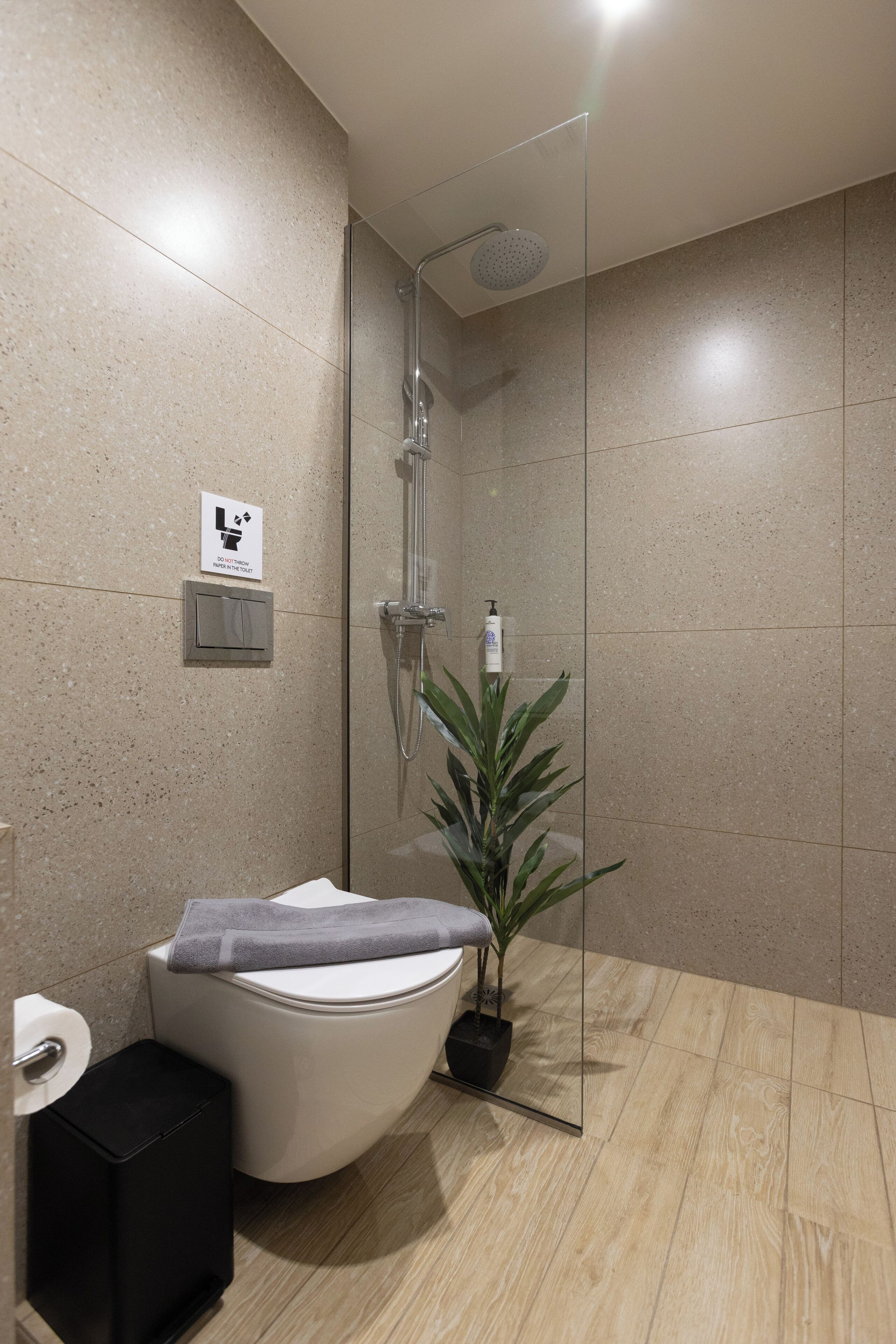 A modern bathroom with a glass-enclosed shower, a toilet, a small trash bin, and a potted plant.