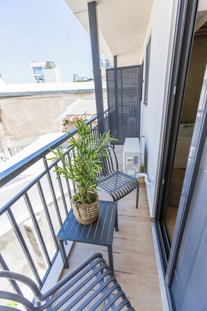 Un balcó amb barana metàl·lica, una planta en test, una taula amb caires i una porta corredissa de vidre que dona a l'interior.