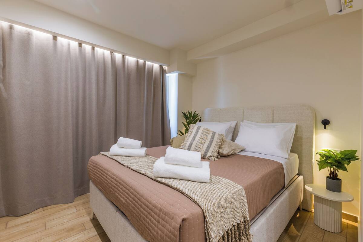 Un lit bien fait, recouvert d’une couverture beige et marron, avec deux oreillers et deux serviettes pliées. Une plante en pot est posée sur une petite table à côté du lit.