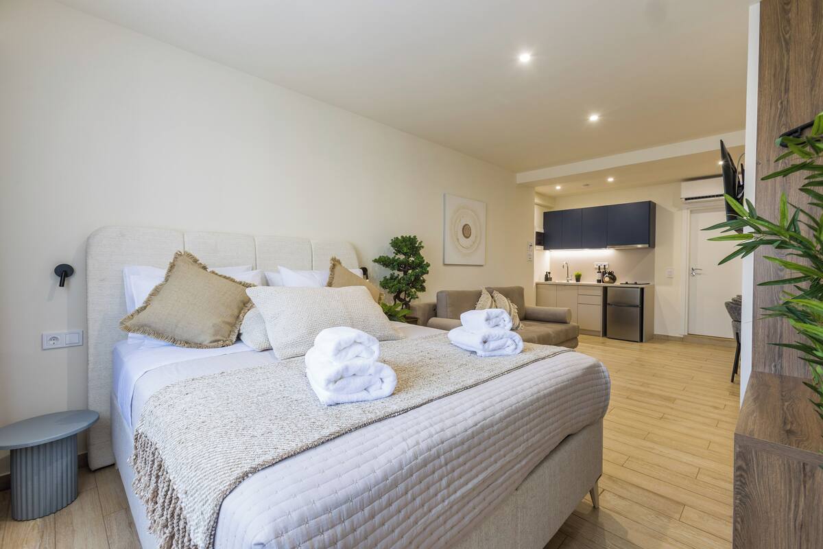 Une chambre d’hôtel moderne avec un grand lit, une petite table ronde, un canapé, un coin cuisine et une plante en pot.