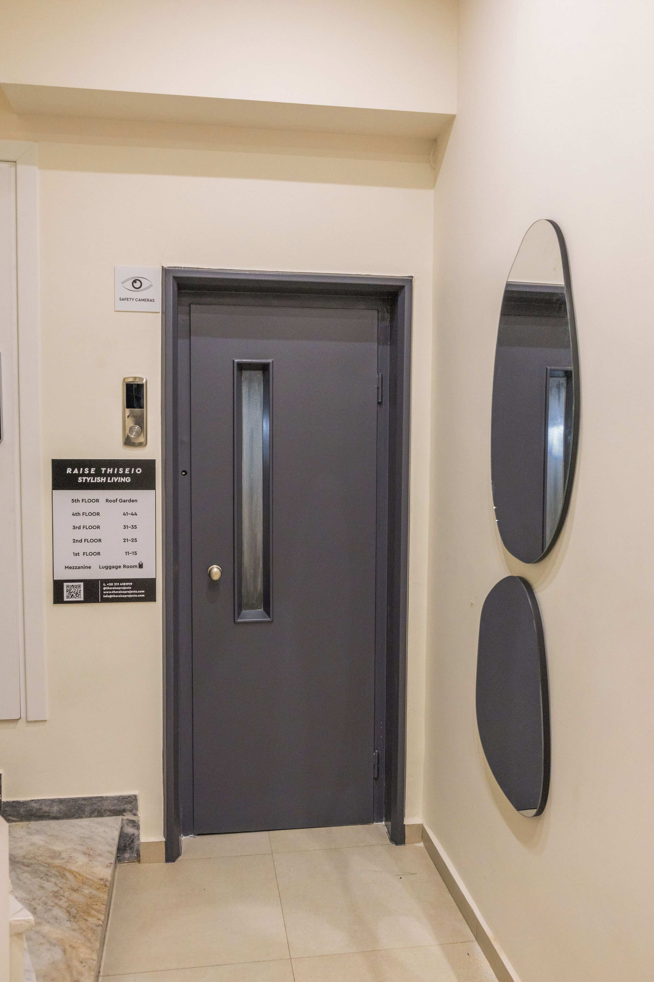A hotel entrance with a dark grey door, a safety camera sign, a directory for room floors, and a wall-mounted mirror.