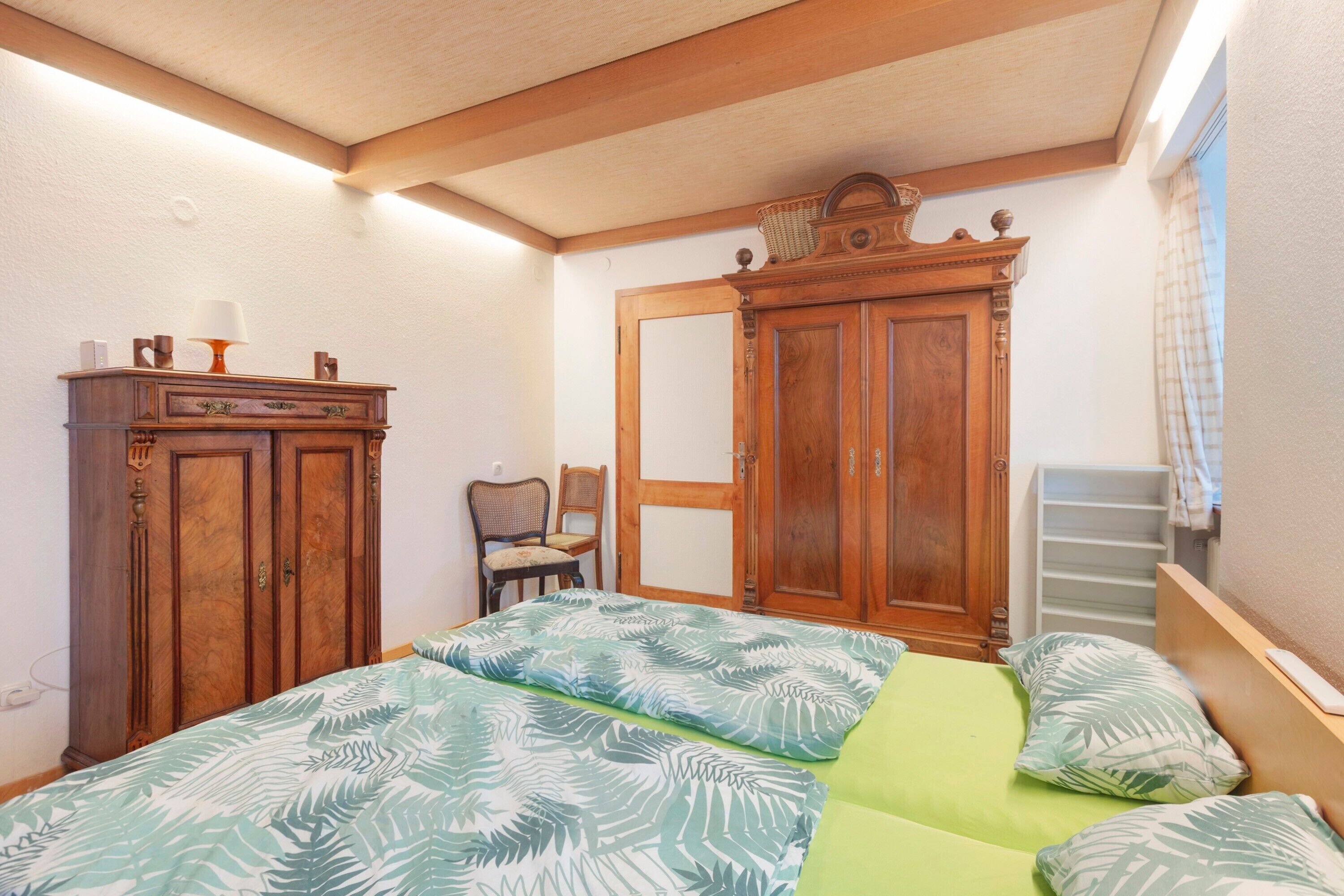 A bedroom with a bed, a wooden wardrobe, a chair, and a window with curtains.