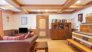Ein gemütliches Wohnzimmer mit einer Couch, einem Holztisch und einer Bank. Es gibt einen Fernseher, eine Holzkommode mit verschiedenen Gegenständen und eine Tür zu einem weiteren Raum.
