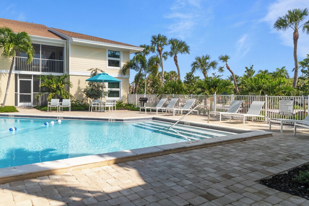 A pool area with lounge chairs and umbrellas, surrounded by palm trees and a building with a balcony.