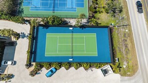 Une vue aérienne d’un court de tennis avec une piscine, entouré de verdure et bordé par une route.