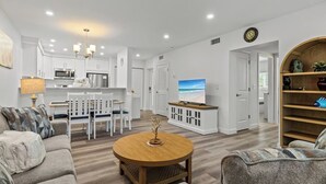 A modern living room with a round wooden coffee table, a sofa, and a TV. There is a kitchen area with white cabinets and a dining table with chairs.