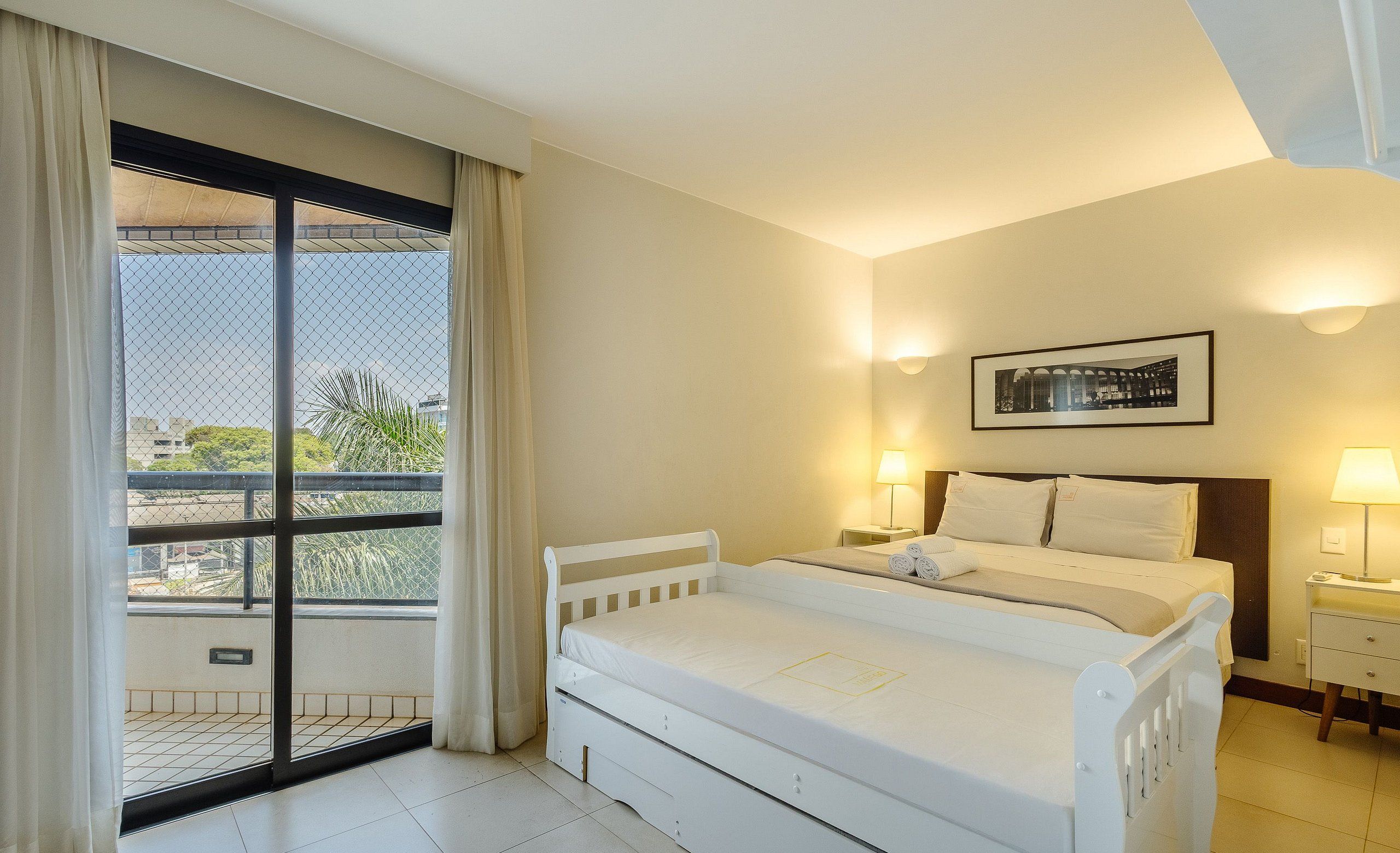 A hotel room with a bed, a nightstand, a lamp, a framed picture on the wall, and a view through a window.