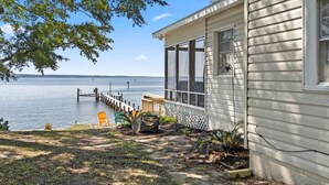 A house with a dock extending into a calm lake, surrounded by greenery. - Ferry Breeze Beach House – NC Coastal Escape (Minnesott Beach)