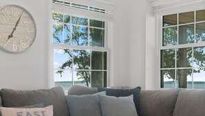 A living room with a couch, a clock, and large windows with a view of trees and water. - Ferry Breeze Beach House – NC Coastal Escape (Minnesott Beach)