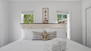 A neatly made bed with white linens, a decorative pillow, and a folded towel. There are two windows with white frames, one of which has a view of greenery outside. - Ferry Breeze Beach House – NC Coastal Escape (Minnesott Beach)
