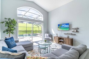 A modern living room with a large glass door leading to an outdoor area, a flat-screen TV mounted on the wall, and a cozy seating arrangement. - 5BR Emerald Island Vacation Home – Game Room, Resort Amenities. Near Theme Parks (Kissimmee)