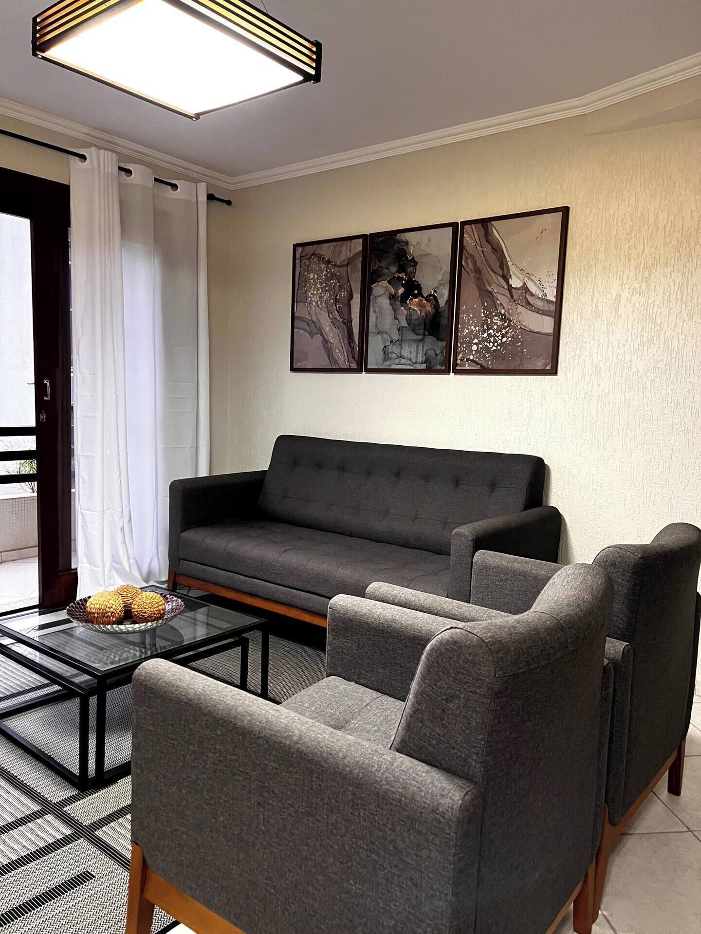 Un salon moderne avec un canapé gris, deux fauteuils, une table basse en verre et des œuvres d’art abstraites accrochées au mur.