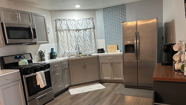 A modern kitchen with stainless steel appliances, light wood flooring, and a built-in brick backsplash.