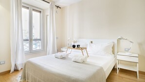A hotel room with a large bed, a bedside table with a lamp, a window with curtains, and a tray with towels and a cup.
