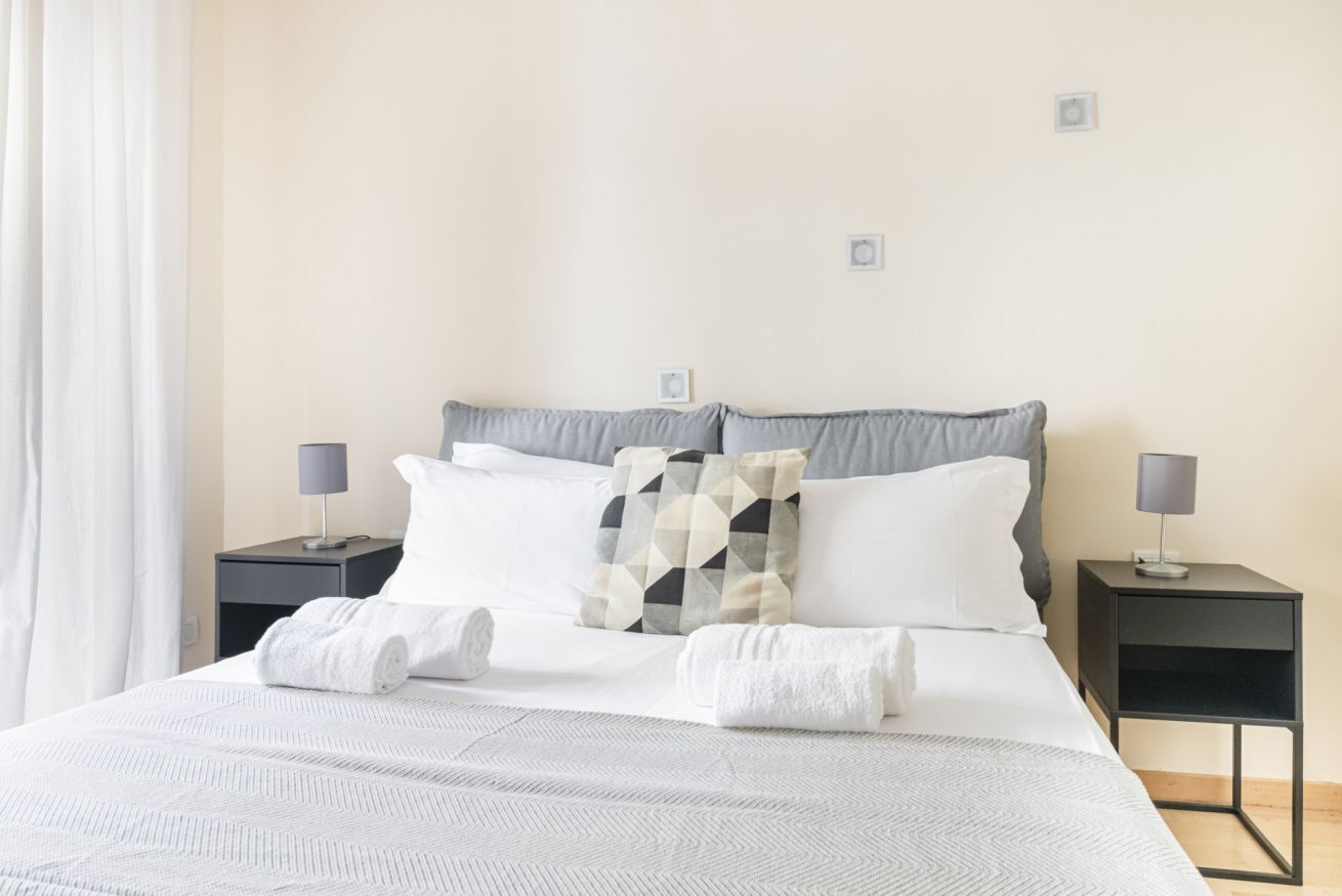 Un lit bien fait, avec du linge de lit blanc et un coussin à motifs géométriques, deux tables de chevet avec des lampes, et un mur sobre orné d’un petit cadre.