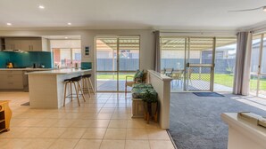 A modern kitchen with a central island, bar stools, and a sliding glass door leading to a patio with outdoor furniture. - Marine Holiday Home - Geographe (Geographe)