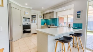 A modern kitchen with a built-in oven, a large island with bar stools, and a view of the backyard through the window. - Marine Holiday Home - Geographe (Geographe)