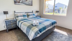 A neatly made bed with a checkered bedspread, two bedside tables with lamps, and a window with a view of a house and yard. - Marine Holiday Home - Geographe (Geographe)