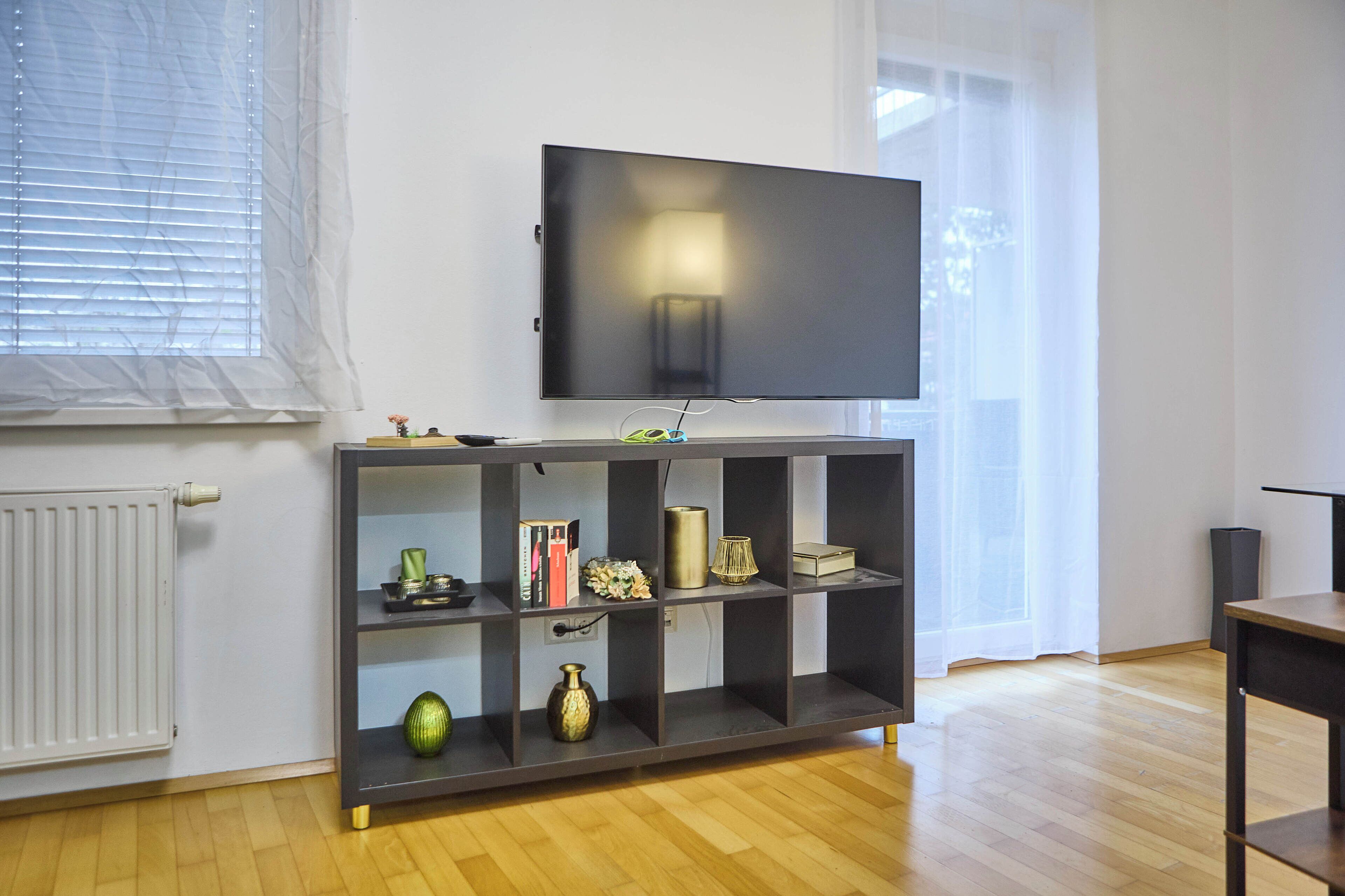 A modern living room with a flat-screen TV mounted on a dark wooden shelf, a radiator, and a window with blinds.