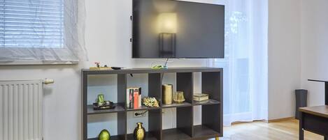 A modern living room with a flat-screen TV mounted on a dark wooden shelf, a radiator, and a window with blinds.