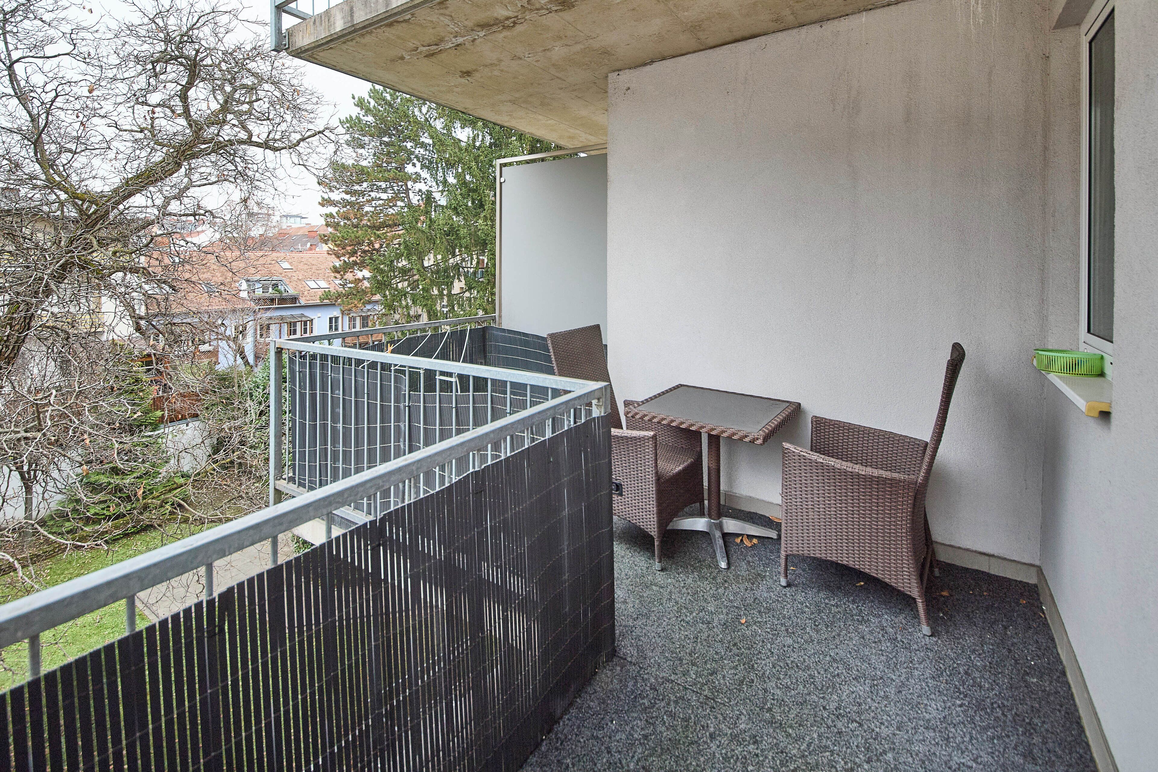 Un balcon avec une table et deux chaises en osier, une clôture en métal et une vue sur les arbres et les bâtiments.