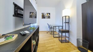 Il s’agit d’une pièce de taille modeste, équipée d’un téléviseur à écran plat, d’un bureau avec un ordinateur, d’une chaise et d’une table. Deux tableaux ornent le mur et une lampe est posée sur la table.